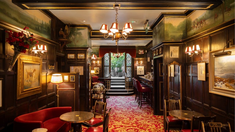 View through an elegant cocktail bar with dark wood panelling, red upholstered seating, framed artwork, and ornate lighting, leading towards a bright glass-roofed winter garden filled with greenery.